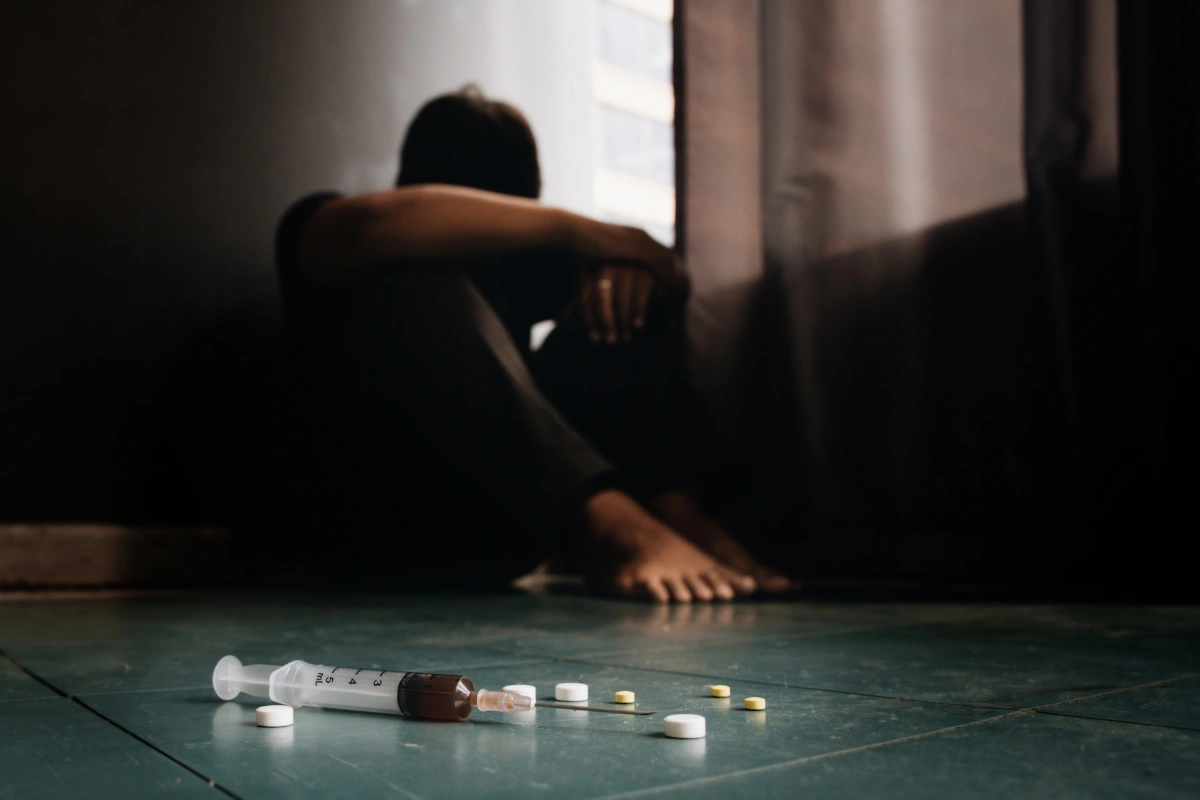 Person sitting alone in the dark with a syringe and pills, showing opioid addiction