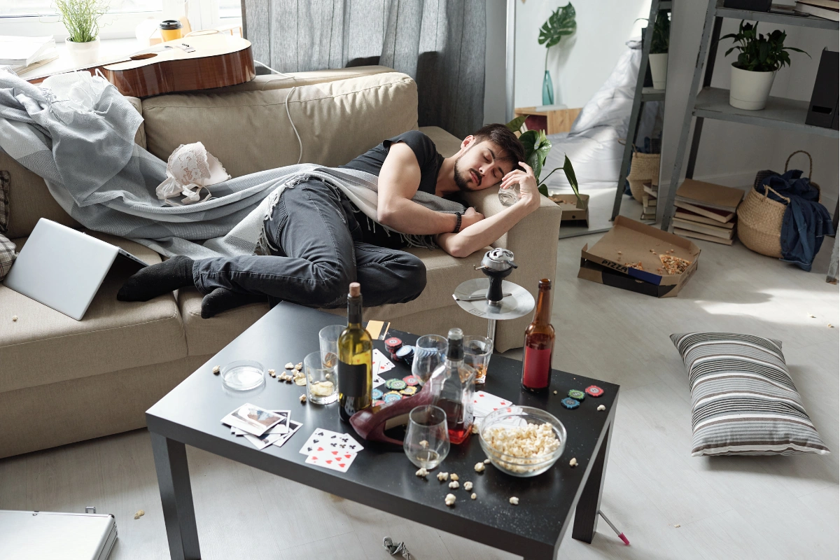 Man asleep on a sofa among empty alcohol bottles after a party