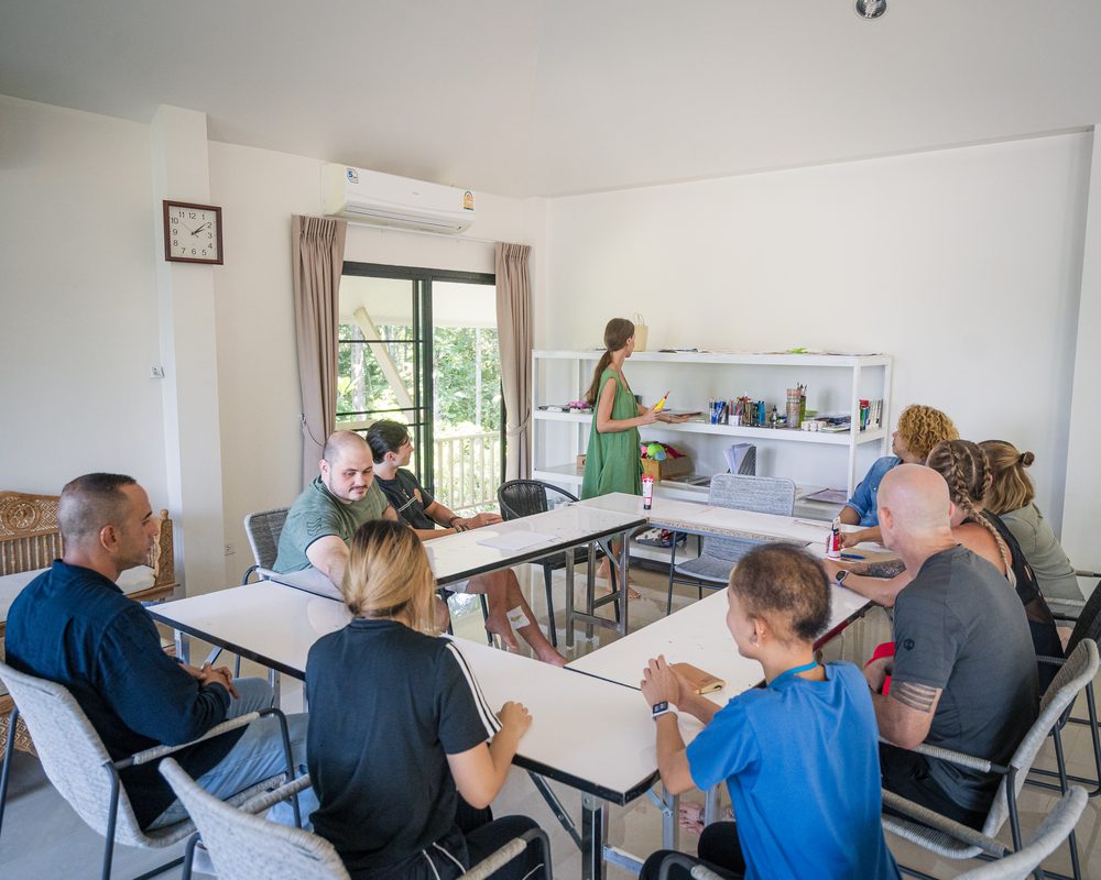 Clients seated around tables in a session room, with a facilitator standing at the materials shelves