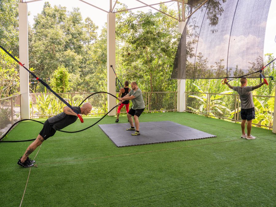 Clients training with battle ropes and TRX straps on the covered outdoor fitness deck, forest beyond the rail