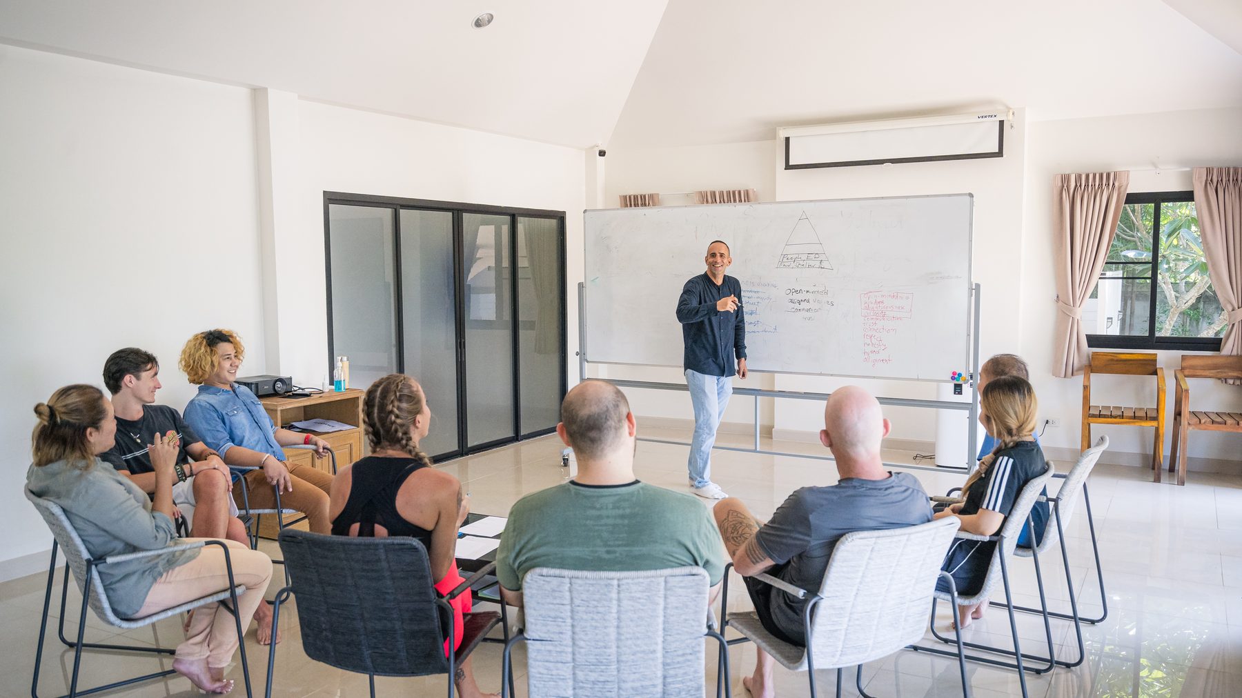 A therapist at a whiteboard leading a group session, eight people seated in a circle of chairs